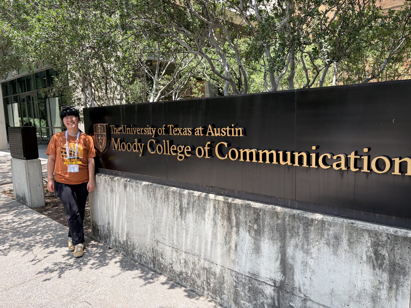 Emily, in a burnt-orange University of Texas shirt, standing and smiling beside the engraved campus sign reading: The University of Texas at Austin, Moody College of Communication.
