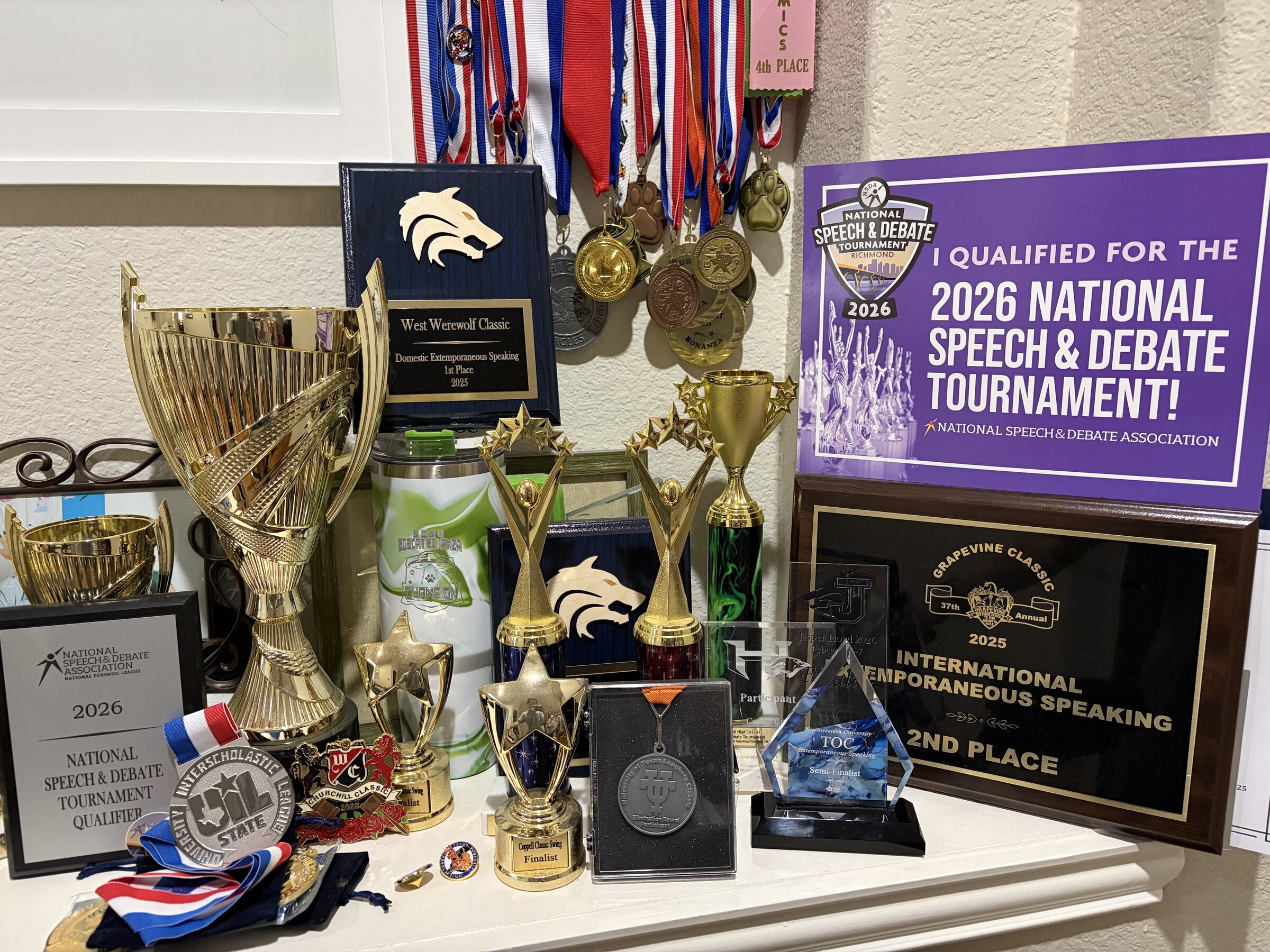 A shelf of speech-and-debate awards: gold trophies and plaques, a blue crystal engraved Extemporaneous Tournament of Champions Semi-Finalist, a silver University Interscholastic League UIL State medal on a red-white-and-blue ribbon, a purple sign reading I qualified for the 2026 National Speech and Debate Tournament, and around two dozen medals on ribbons hanging behind them.